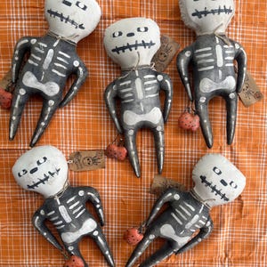 May include: Five handmade fabric skeleton dolls with painted details. Each doll is holding a small orange fabric pumpkin. The dolls are arranged on a checkered orange and white fabric.