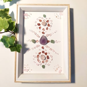 May include: A framed crystal grid with a white background. The grid is made of small pink, green, and red crystals. A larger amethyst crystal is in the center of the grid.