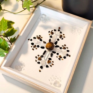May include: A rectangular wooden shadow box frame containing a white backing with a central tiger's eye sphere surrounded by a cross-shaped arrangement of clear quartz points, small black stones, and brown tiger's eye chips on a white surface.