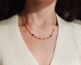 May include: A delicate gold chain necklace adorned with small, colorful beads. The necklace features a mix of pink, red, and gold-colored beads, creating a subtle and elegant design. The necklace is worn on a person.