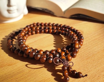 Puede incluir: Un collar de cuentas de mala de madera marrón con un borla. Las cuentas son redondas y de color marrón oscuro. La borla está hecha de cuerda marrón y tiene un nudo en el extremo.