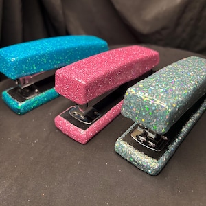 Może przedstawiać: Trzy zszywacze pokryte brokatem w kolorach turkusowym, różowym i iryzującym. Zszywacze mają metalową, srebrną podstawę i są ułożone na ciemnej powierzchni. Artykuły biurowe.