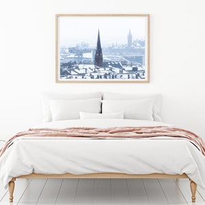May include: A framed photograph of a snowy cityscape with a tall church steeple in the centre. The photo is hanging on a white wall above a white bed with a pink throw blanket.