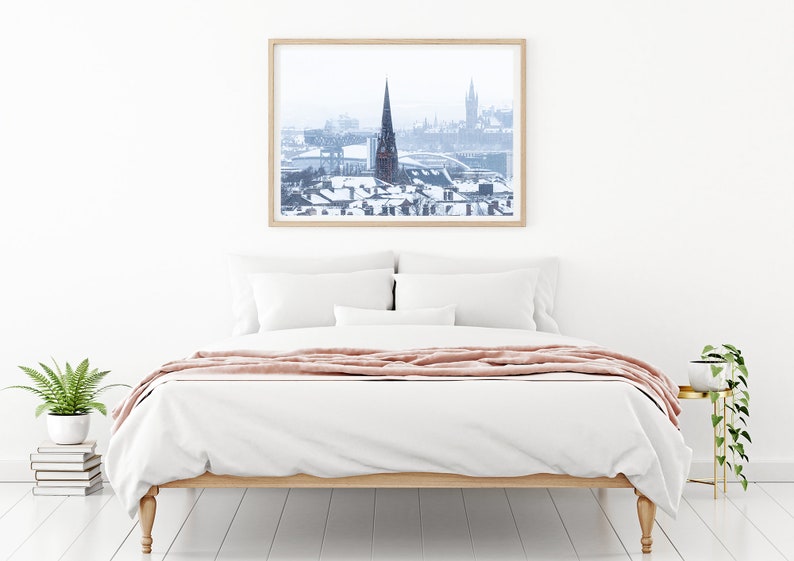 May include: A framed photograph of a snowy cityscape with a tall church steeple in the centre. The photo is hanging on a white wall above a white bed with a pink throw blanket.