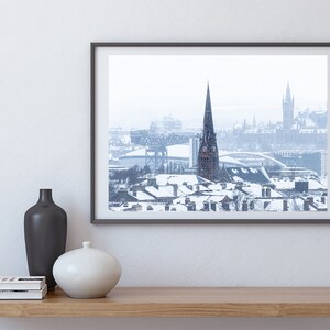 May include: A framed photograph of a snowy cityscape with a tall church spire in the centre. The photo is in a black frame and is hanging on a white wall.
