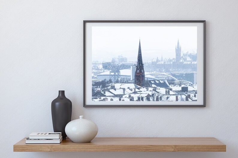 May include: A framed photograph of a snowy cityscape with a tall church spire in the centre. The photo is in a black frame and is hanging on a white wall.