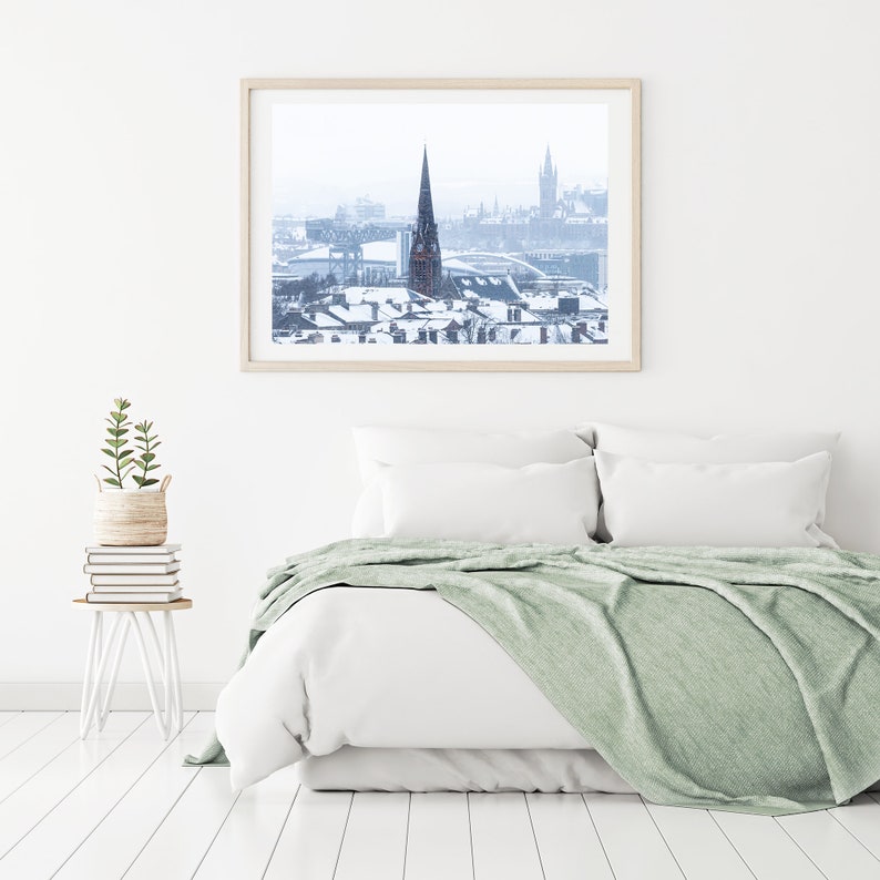 May include: A framed photograph of a snowy cityscape with a tall church steeple in the centre. The photo is hanging on a white wall above a white bed with a green throw blanket.