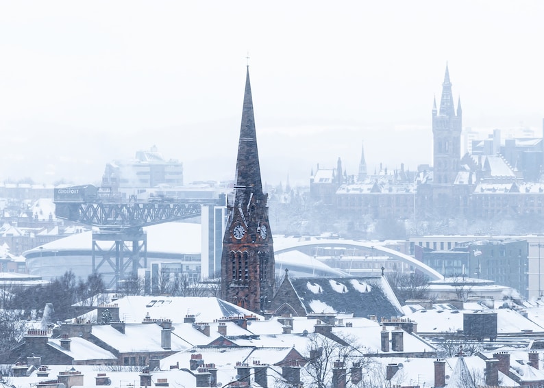 May include: A snowy cityscape with a tall, pointed church spire in the foreground. The church is brick with a clock face and a spire. The background shows a cityscape with a large building with a tower and other buildings covered in snow.