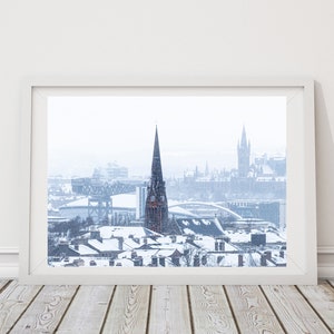 May include: A snowy cityscape with a tall church steeple in the centre. The church is surrounded by other buildings and a bridge in the distance. The sky is overcast and the snow is falling.