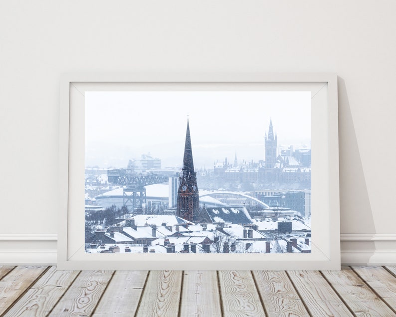 May include: A snowy cityscape with a tall church steeple in the centre. The church is surrounded by other buildings and a bridge in the distance. The sky is overcast and the snow is falling.