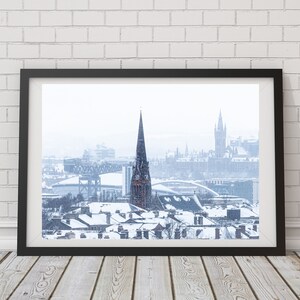 May include: A snowy cityscape with a tall church steeple in the centre, a bridge in the background, and snow-covered rooftops in the foreground.