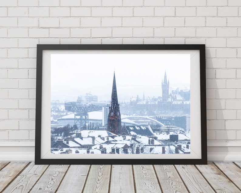 May include: A snowy cityscape with a tall church steeple in the centre, a bridge in the background, and snow-covered rooftops in the foreground.