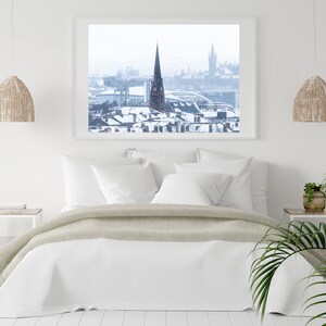 May include: A white bedroom with a bed, two bedside tables, and a framed photograph of a snowy city skyline. The photograph shows a tall church steeple in the centre of the image.