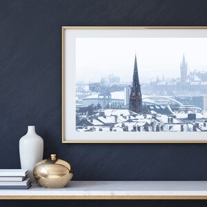 May include: A framed photograph of a snowy cityscape with a tall church spire in the centre. The city is covered in snow and the sky is overcast.