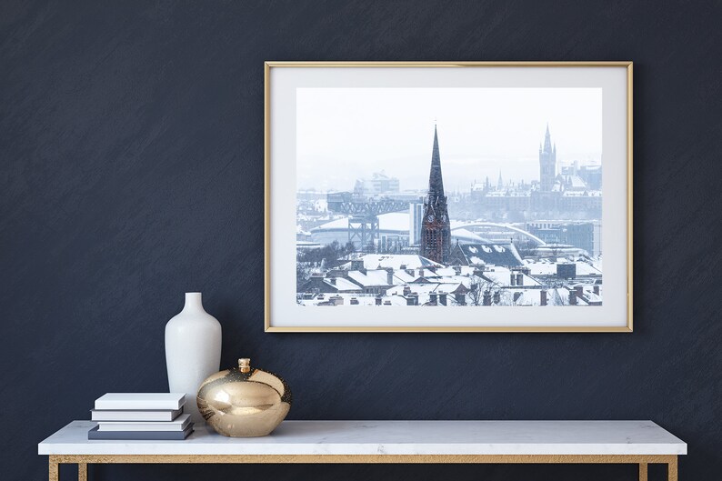 May include: A framed photograph of a snowy cityscape with a tall church spire in the centre. The city is covered in snow and the sky is overcast.