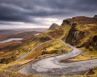 May include: Scenic landscape featuring a winding road through rugged, grassy hills under a dramatic, cloudy sky. The road curves sharply, offering views of a distant lake and the surrounding terrain. The color palette is dominated by greens, browns, and grays.