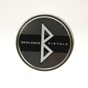 Könnte beinhalten: Ein runder Metallbehälter mit den Worten "BERLOQUE PISTOLE" in Weiß auf einem schwarzen Streifen. Das Design umfasst ein großes, stilisiertes weißes "B" und den Text "Die kleinste Pistole der Welt" in einem kreisförmigen Muster.