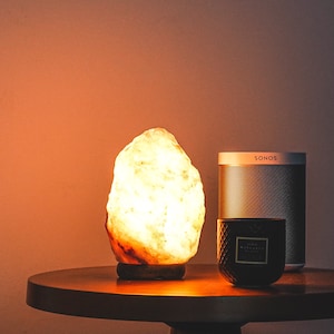 May include: A warm, glowing Himalayan salt lamp sits on a wooden table next to a black candle and a silver speaker labeled "SONOS".