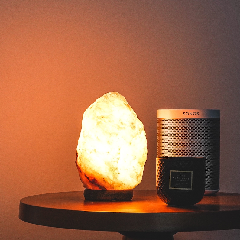 May include: A warm, glowing Himalayan salt lamp sits on a wooden table next to a black candle and a silver speaker labeled "SONOS".