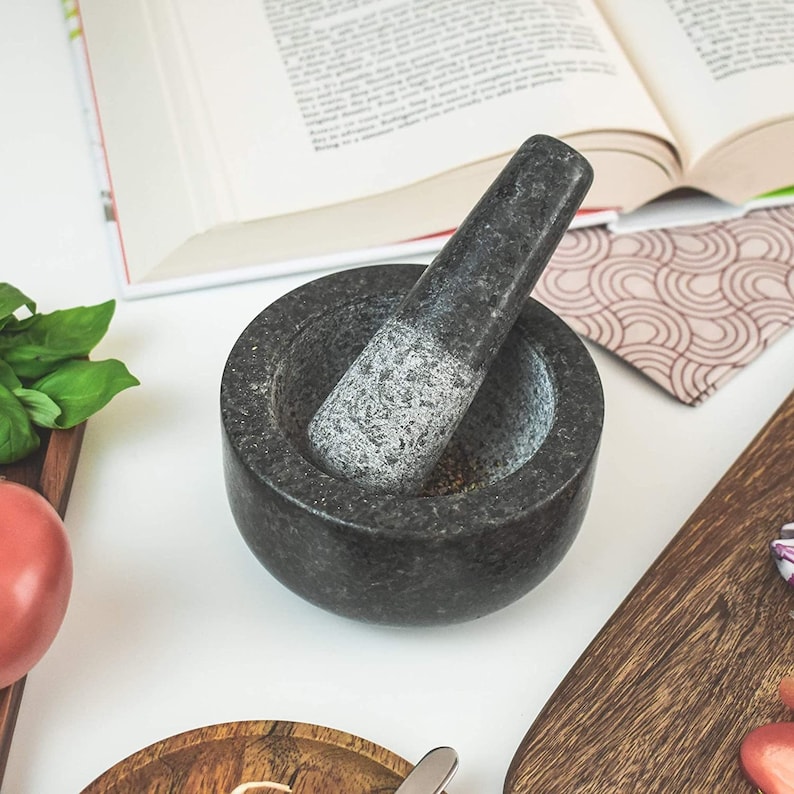 May include: A black granite mortar and pestle set. The mortar is round and has a textured surface. The pestle is long and has a smooth surface.