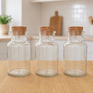 May include: Three clear glass jars with cork lids are displayed on a light wood surface. The jars have a cylindrical shape with a wider base and a narrow neck. The cork lids are light brown. The background shows a kitchen setting.