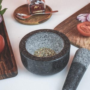 May include: A gray granite mortar and pestle set with a small amount of dried herbs inside the mortar. The mortar and pestle are on a white surface with a wooden cutting board and a small glass jar in the background.