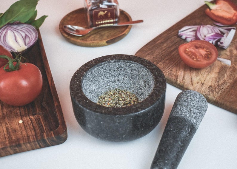 May include: A gray granite mortar and pestle set with a small amount of dried herbs inside the mortar. The mortar and pestle are on a white surface with a wooden cutting board and a small glass jar in the background.
