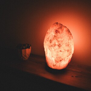 May include: A large, pink Himalayan salt lamp glows with a warm orange light, casting a soft glow on the wooden surface it sits on.