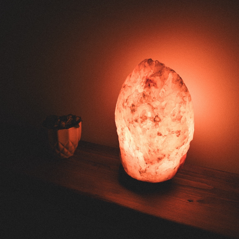 May include: A large, pink Himalayan salt lamp glows with a warm orange light, casting a soft glow on the wooden surface it sits on.