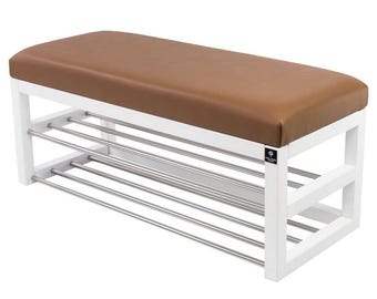 Op de afbeelding: Een rechthoekige schoenenbank met een wit houten frame en een gladde bruine gestoffeerde zitting. De basis bevat twee niveaus van horizontale zilverkleurige metalen stangen voor opslag. Een klein zwart label met witte tekst is bevestigd aan het zijframe.