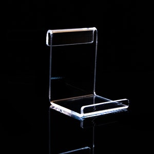 May include: A clear acrylic display stand for showcasing small items. The stand has a rectangular base and a curved back.