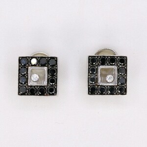 Peut inclure: Une paire de boucles d'oreilles carrées. Chaque boucle d'oreille présente un cadre carré de pierres noires entourant un centre clair. Le centre contient une seule pierre blanche. Les boucles d'oreilles sont posées sur un fond blanc.