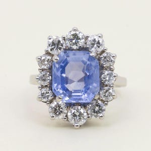 May include: A silver ring featuring a large, light blue, rectangular gemstone surrounded by a halo of smaller, round, clear gemstones. The ring's design is classic and elegant, suitable for special occasions.