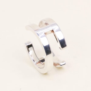 May include: A silver-colored ear cuff with a modern, minimalist design. The cuff features a double-banded structure with a rectangular opening in the center. The jewelry is made of a shiny, reflective metal.