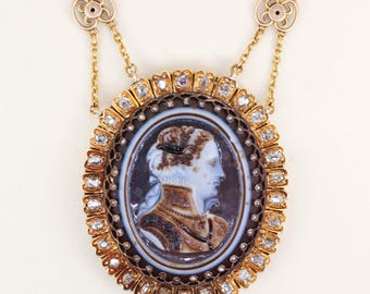 Puede incluir: Un collar de oro con un colgante ovalado que presenta un retrato de camafeo de una mujer de perfil. El camafeo está rodeado de pequeños diamantes y un marco marrón oscuro. El collar tiene delicadas cadenas de oro y detalles florales decorativos.