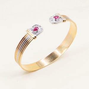 Può includere: Un bracciale a polsino color oro con dettagli argento e oro. Ogni estremità presenta un elemento decorativo con una gemma rossa e piccole pietre trasparenti. Il bracciale ha un design semplice ed elegante.