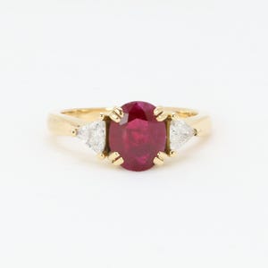 May include: A gold ring featuring a large oval ruby center stone flanked by two triangular diamonds. The ring's band is a simple, polished gold, and the gemstones are set against a white background.