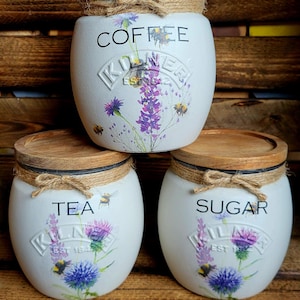 May include: Three white ceramic canisters with wooden lids and twine accents. The canisters are decorated with floral designs and the words "Coffee", "Tea", and "Sugar".