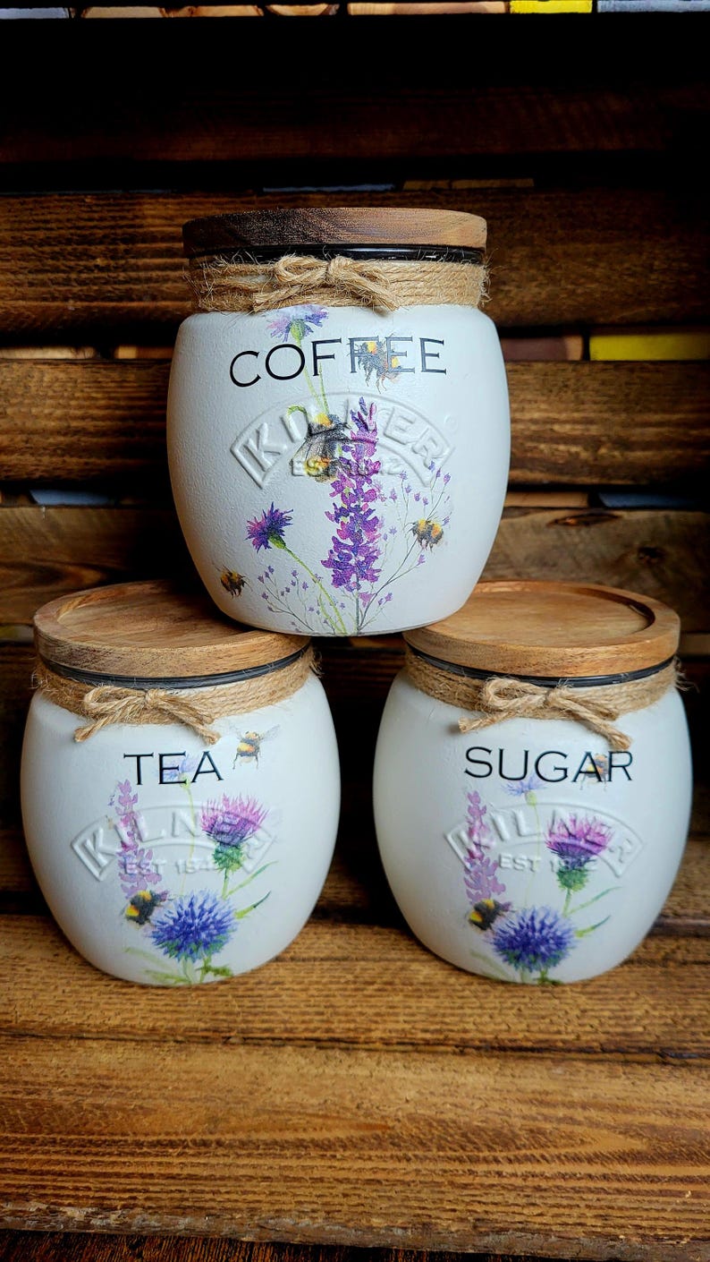 May include: Three white ceramic canisters with wooden lids and twine accents. The canisters are decorated with floral designs and the words "Coffee", "Tea", and "Sugar".