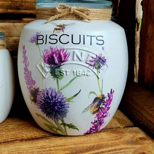 May include: White ceramic biscuit jar with a glass lid and a floral design featuring purple flowers, green leaves, and yellow bumblebees. The jar has the text "BISCUITS" printed in black on the front and "EST 1842" printed in black below the flowers.