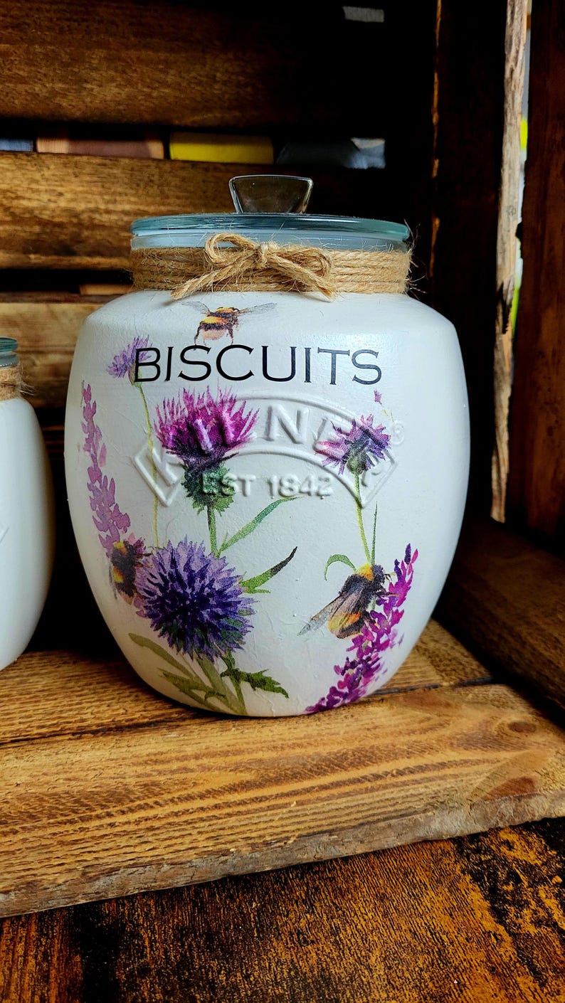 May include: White ceramic biscuit jar with a glass lid and a floral design featuring purple flowers, green leaves, and yellow bumblebees. The jar has the text "BISCUITS" printed in black on the front and "EST 1842" printed in black below the flowers.
