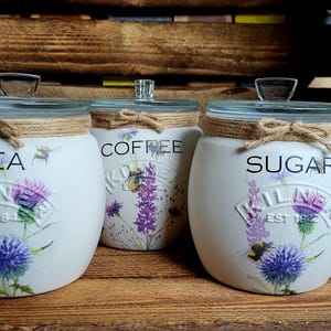 May include: Three white ceramic canisters with glass lids and twine accents. The canisters are decorated with floral designs and the words "Tea", "Coffee", and "Sugar".