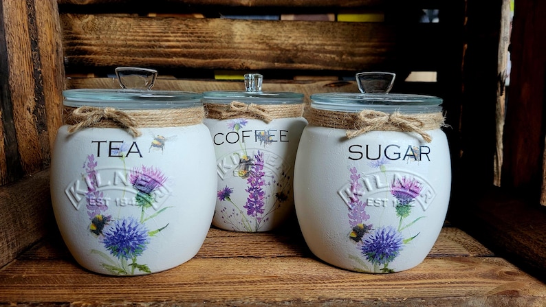 May include: Three white ceramic canisters with glass lids and twine accents. The canisters are decorated with floral designs and the words "Tea", "Coffee", and "Sugar".