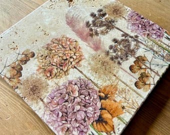 May include: A square travertine stone tile featuring a printed botanical design of dried flowers, including purple hydrangea blossoms, pampas grass, and various seed heads in shades of brown, tan, and muted pink against a porous beige background.