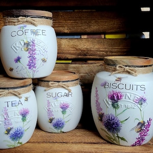 May include: Four white ceramic canisters with wooden lids and floral designs. The canisters are labelled "Coffee", "Tea", "Sugar", and "Biscuits".