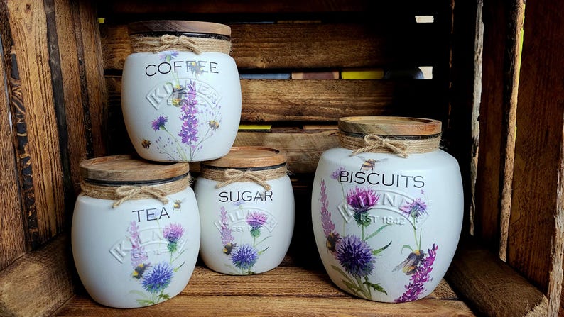 May include: Four white ceramic canisters with wooden lids and floral designs. The canisters are labeled "Coffee", "Tea", "Sugar", and "Biscuits".