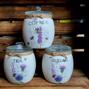 May include: Three white ceramic canisters with floral designs and lids. The canisters are labeled "Coffee", "Tea", and "Sugar".