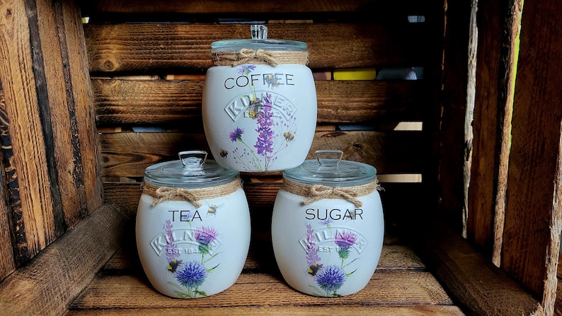 May include: Three white ceramic canisters with floral designs and lids. The canisters are labeled "Coffee", "Tea", and "Sugar".