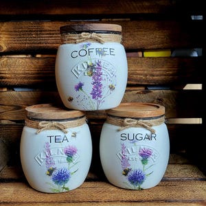 May include: Three white ceramic canisters with wooden lids and floral designs. The canisters are labeled "Coffee", "Tea", and "Sugar".