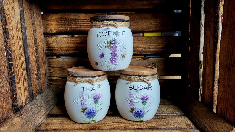May include: Three white ceramic canisters with wooden lids and floral designs. The canisters are labeled "Coffee", "Tea", and "Sugar".